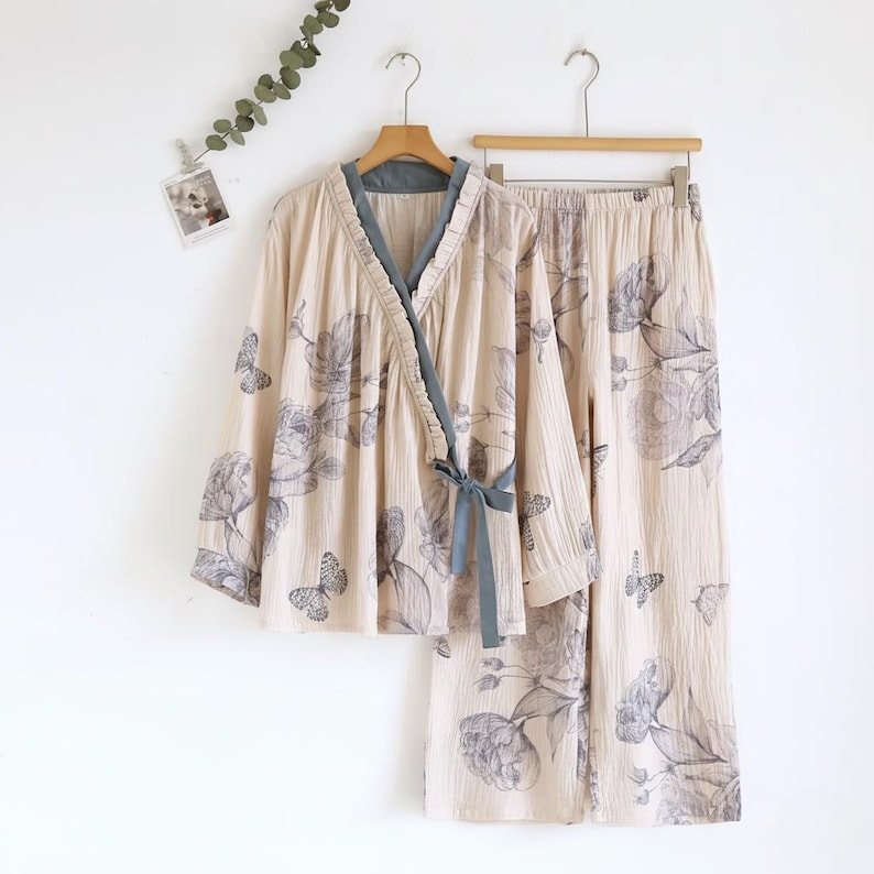 May include: A beige pajama set with a floral and butterfly design. The wrap-style top has a grey trim and tie. The matching trousers are wide-legged. The set is displayed on a wooden hanger.