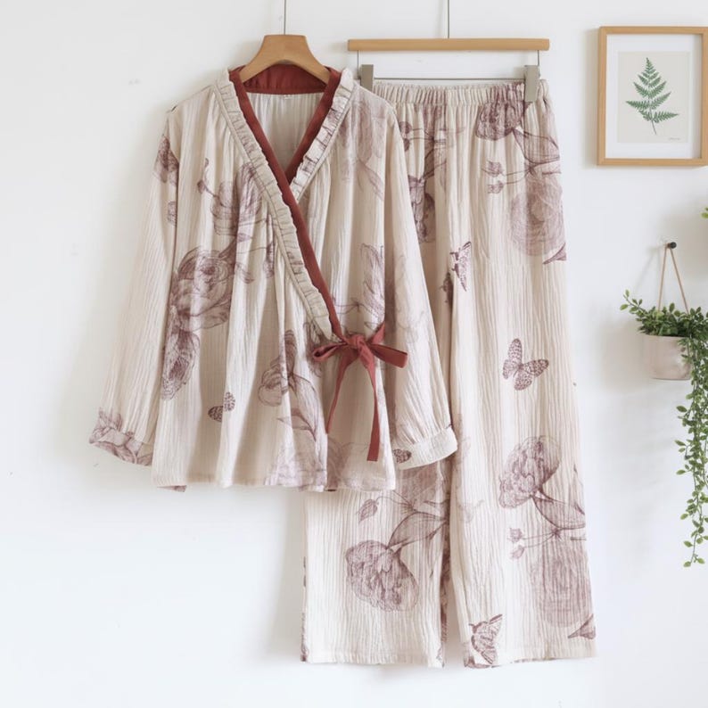 May include: Cream-coloured two-piece pyjama set with a floral and butterfly pattern. The top has a wrap-style design with a brown trim and tie. The trousers are wide-legged. The set is hanging on a wooden hanger.