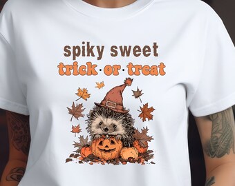May include: White t-shirt with a cute cartoon hedgehog wearing a witch hat and holding a pumpkin. The text "spiky sweet trick or treat" is printed on the shirt.