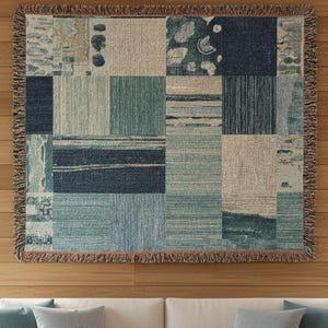 May include: A woven throw blanket with a patchwork design in shades of blue, green, and beige hangs on a wooden wall. The blanket has a fringed edge. A white sofa with blue pillows sits below the blanket. A wooden coffee table and a vase of dried flowers are in front of the sofa.