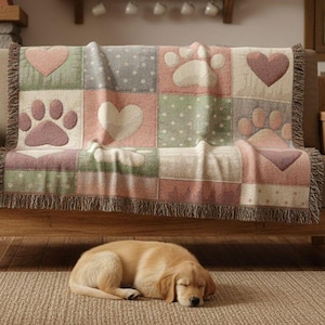 May include: A throw blanket with a patchwork design featuring hearts and paw prints in pink, green, and gray tones. The blanket is draped over a wooden sofa. A sleeping golden retriever dog rests on a rug in front of the sofa.