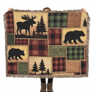 Peut inclure: Une couverture tissée avec un motif patchwork représentant des silhouettes noires d'orignaux, d'ours et de pins. La couverture a une bordure à franges et une palette de couleurs beige, rouge, vert et marron.