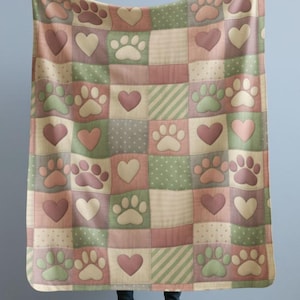 May include: A patchwork blanket featuring a repeating pattern of paw prints and hearts in shades of pink, green, and cream. The blanket is held up, showcasing its design and soft color palette. The squares are approximately 20 cm.