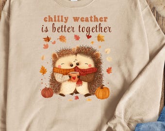 May include: Tan sweatshirt with a graphic of two hedgehogs holding a mug of hot cocoa. The text "chilly weather is better together" is printed above the hedgehogs.