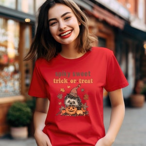 May include: Red t-shirt with a cute cartoon hedgehog wearing a witch hat and holding a jack-o-lantern. The text "spiky sweet trick or treat" is printed on the shirt.