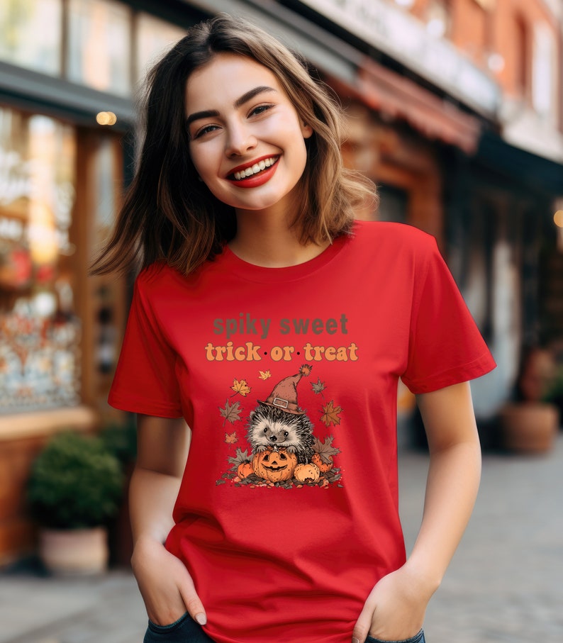 May include: Red t-shirt with a cute cartoon hedgehog wearing a witch hat and holding a jack-o-lantern. The text "spiky sweet trick or treat" is printed on the shirt.