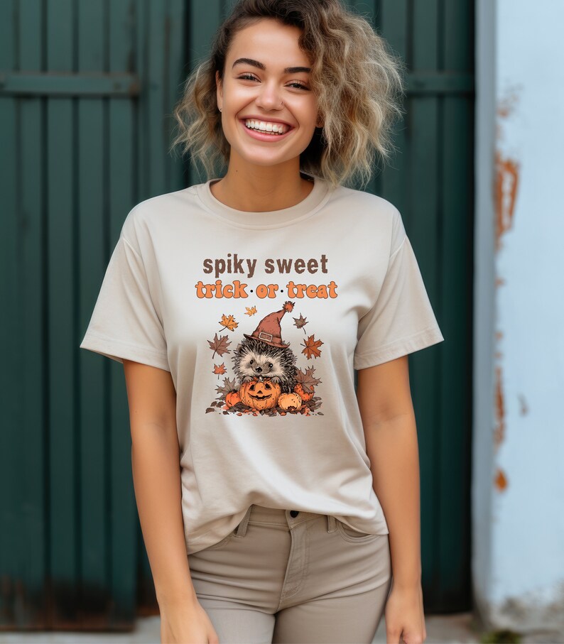 May include: A beige t-shirt with a cute hedgehog wearing a witch hat and holding a pumpkin. The text "spiky sweet trick or treat" is printed on the shirt.