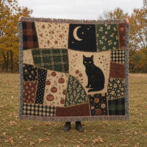 Könnte beinhalten: Eine gewebte Wohndecke mit Patchwork-Design, das verschiedene Muster wie Karos, Blumen und Kürbisse zeigt. Eine schwarze Katze sitzt mittig auf cremefarbenem Grund, ergänzt durch eine Mondsichel und Sterne in der oberen Ecke.
