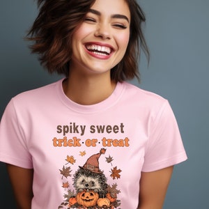 May include: Pink t-shirt with a graphic of a hedgehog wearing a witch hat and holding a pumpkin. The text "spiky sweet trick or treat" is printed on the shirt.