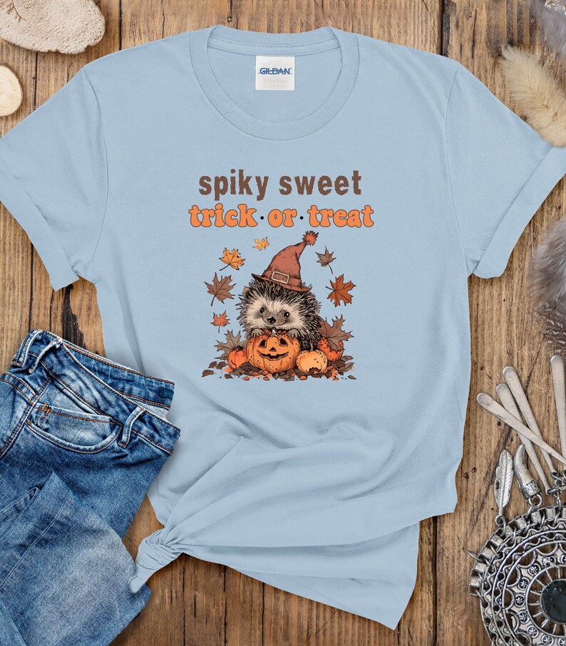 May include: Light blue t-shirt with a cartoon hedgehog wearing a witch's hat and holding a jack-o'-lantern. The text "spiky sweet trick or treat" is printed on the shirt.