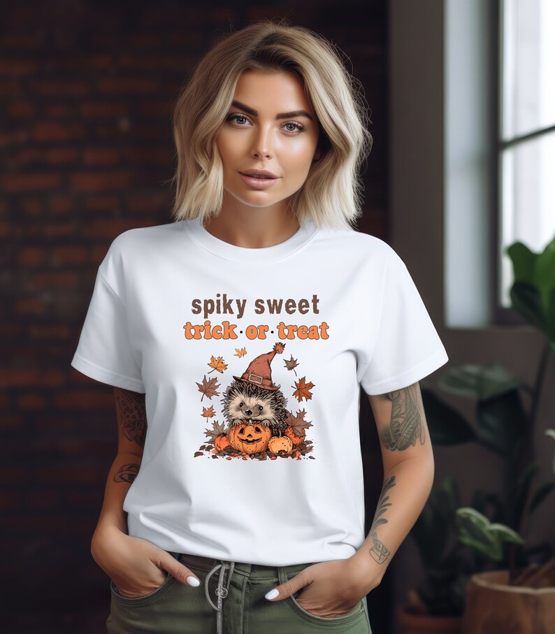 May include: White t-shirt with a cute cartoon hedgehog wearing a witch hat and holding a pumpkin. The text "spiky sweet trick or treat" is printed on the shirt.