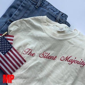 May include: A white t-shirt with the text "The Silent Majority" embroidered in red. The shirt is folded and laid on top of a pair of blue jeans.