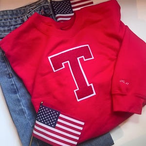 May include: A red sweatshirt with a large white-outlined letter "T" on the front. The sleeve has "No. 47" embroidered in white. The sweatshirt is paired with blue jeans and small American flags.