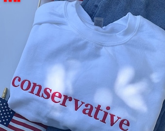 Puede incluir: Sudadera blanca con la palabra "conservador" bordada en rojo en la parte delantera.