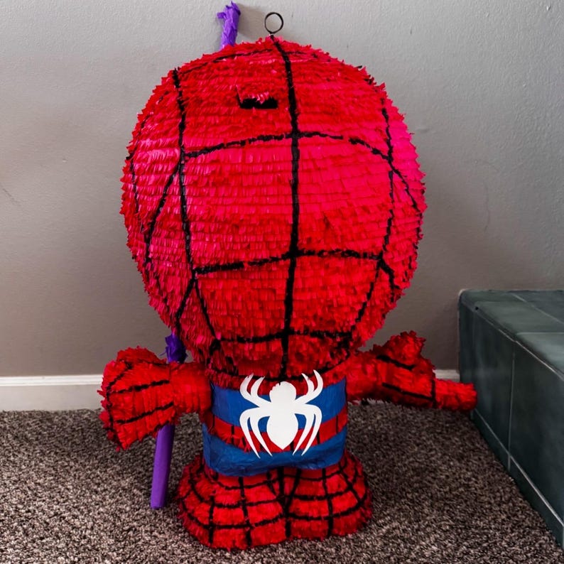 Puede incluir: Una pi&ntilde;ata de Spider-Man roja con detalles de telara&ntilde;a negra. La pi&ntilde;ata tiene un cintur&oacute;n azul y blanco con un emblema de ara&ntilde;a. Un palo morado est&aacute; unido a la pi&ntilde;ata.