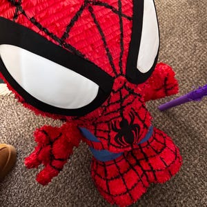 Puede incluir: Una pi&ntilde;ata roja de Spider-Man con detalles de telara&ntilde;a negra y grandes formas de ojos blancos. La pi&ntilde;ata presenta un emblema de ara&ntilde;a negra en el pecho y una cintura azul. La pi&ntilde;ata est&aacute; hecha de papel crep&eacute; rojo.