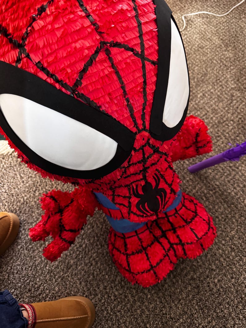 Puede incluir: Una pi&ntilde;ata roja de Spider-Man con detalles de telara&ntilde;a negra y grandes formas de ojos blancos. La pi&ntilde;ata presenta un emblema de ara&ntilde;a negra en el pecho y una cintura azul. La pi&ntilde;ata est&aacute; hecha de papel crep&eacute; rojo.