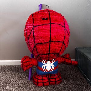 Puede incluir: Una pi&ntilde;ata roja de Spider-Man con l&iacute;neas de telara&ntilde;a negras, un emblema de ara&ntilde;a azul y blanco y brazos extendidos. La pi&ntilde;ata tiene un lazo colgante morado y est&aacute; hecha de papel crep&eacute;. Dise&ntilde;ada para una fiesta.