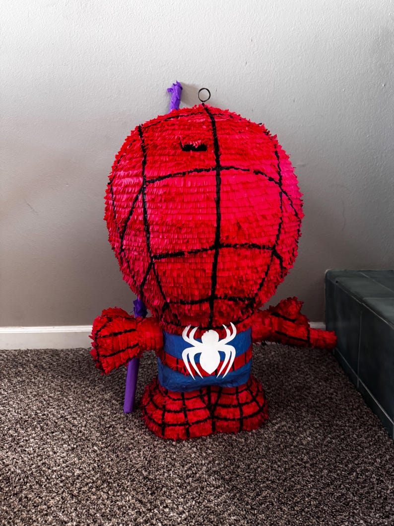 Puede incluir: Una pi&ntilde;ata roja de Spider-Man con l&iacute;neas de telara&ntilde;a negras, un emblema de ara&ntilde;a azul y blanco y brazos extendidos. La pi&ntilde;ata tiene un lazo colgante morado y est&aacute; hecha de papel crep&eacute;. Dise&ntilde;ada para una fiesta.