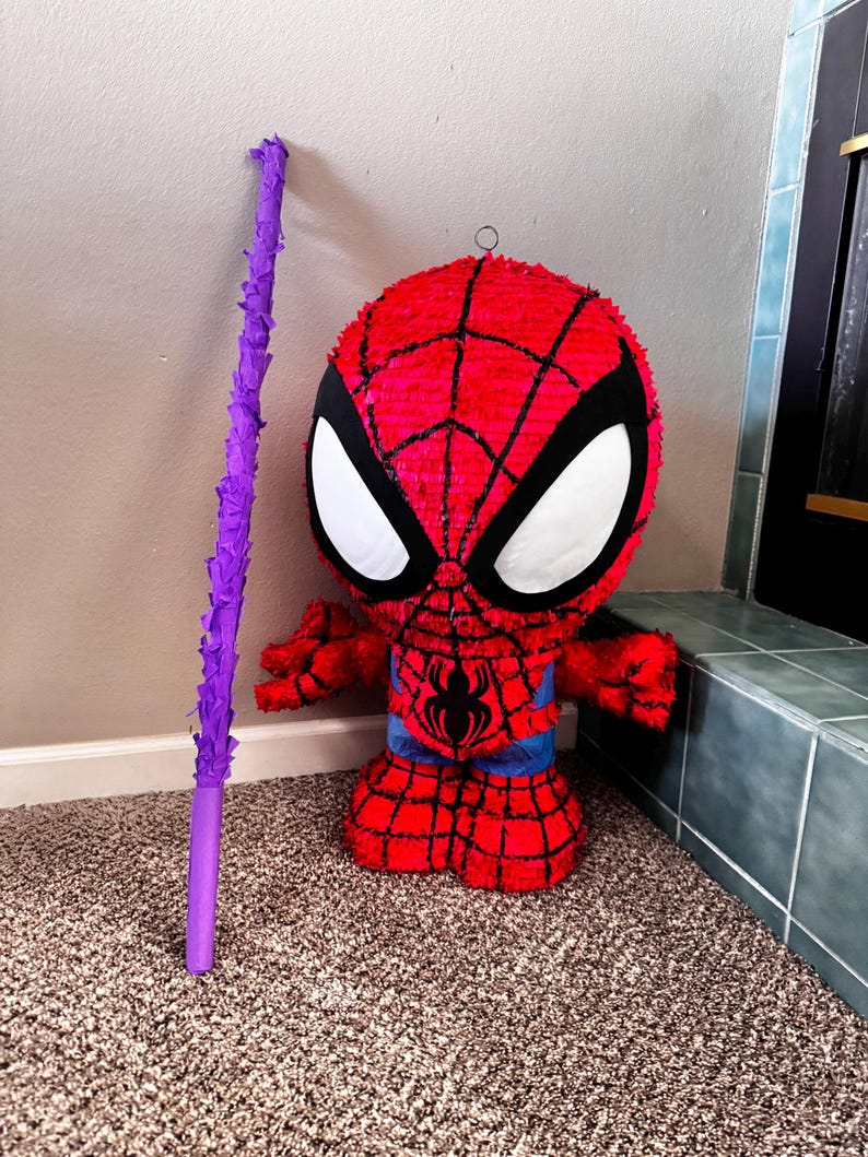 Puede incluir: Una pi&ntilde;ata de Spider-Man roja y negra con recortes blancos para los ojos y una cintura azul. Un palo morado est&aacute; apoyado contra la pared junto a la pi&ntilde;ata. La pi&ntilde;ata est&aacute; hecha de papel crep&eacute; rojo y l&iacute;neas negras.