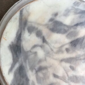 Puede incluir: Un plato redondo, blanquecino, con un patr&oacute;n veteado gris y blanco. El plato tiene un borde elevado y una textura ligeramente rugosa. El dise&ntilde;o se asemeja a una pintura de acuarela.