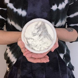 Puede incluir: Un plato redondo de cer&aacute;mica blanca con un dise&ntilde;o abstracto gris y blanco. El plato es sostenido por una persona que lleva una rebeca te&ntilde;ida con tinte anudado en blanco y negro y un vestido azul oscuro.