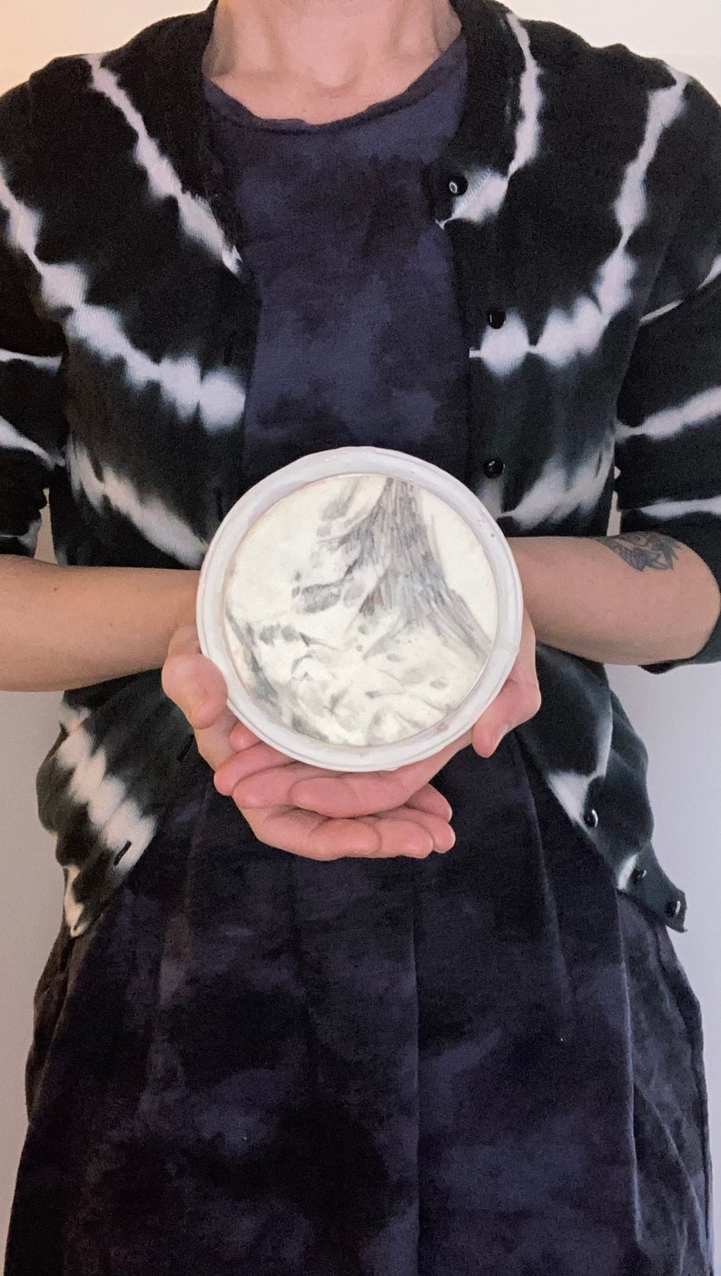 Puede incluir: Un plato redondo de cer&aacute;mica blanca con un dise&ntilde;o abstracto gris y blanco. El plato es sostenido por una persona que lleva una rebeca te&ntilde;ida con tinte anudado en blanco y negro y un vestido azul oscuro.