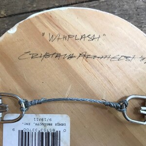 Puede incluir: Parte trasera de una obra de arte de madera con un sistema para colgar con alambre. El t&iacute;tulo manuscrito "WHIPLASH" y la firma del artista son visibles. Tambi&eacute;n hay una etiqueta con un c&oacute;digo de barras y una fecha.