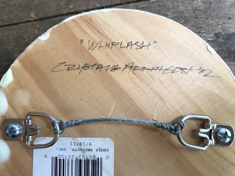 Puede incluir: Parte trasera de una obra de arte de madera con un sistema para colgar con alambre. El t&iacute;tulo manuscrito "WHIPLASH" y la firma del artista son visibles. Tambi&eacute;n hay una etiqueta con un c&oacute;digo de barras y una fecha.