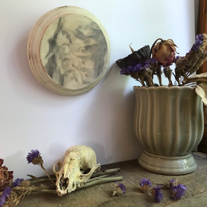 Puede incluir: Un adorno de pared redondo, blanquecino, con una imagen descolorida en escala de grises. Una peque&ntilde;a calavera de animal y un jarr&oacute;n con flores secas se encuentran sobre una superficie de madera. El jarr&oacute;n es de color verde claro y tiene un dise&ntilde;o acanalado. Flores moradas secas est&aacute;n esparcidas alrededor de la calavera.