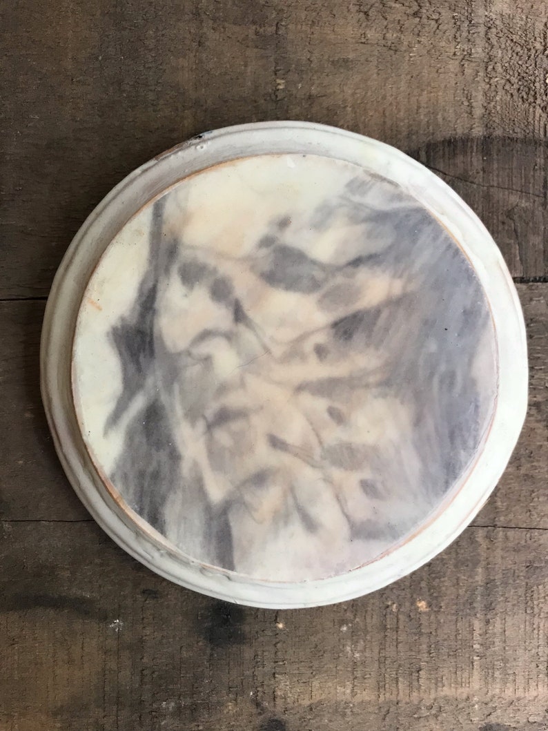 Puede incluir: Un plato decorativo redondo con un dise&ntilde;o veteado en tonos grises y crema. Tiene un borde blanco en relieve y una superficie ligeramente texturizada. Ideal para servir o exhibir, con un di&aacute;metro de unos 25 cm.