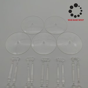 May include: Five clear plastic round bases and six clear plastic supports for miniature figures. The bases are approximately 3 inches in diameter. The supports are approximately 2 inches tall. The text "WAR BASE WEST" is visible in the upper right corner of the image.