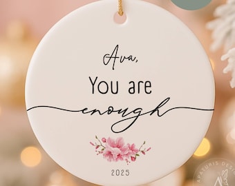 May include: A white ceramic ornament with the text "Ava, You are enough" in black script, accented with pink floral illustrations and the year 2025. A gold string is attached for hanging.