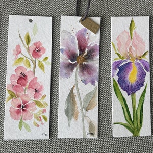 Op de afbeelding: Vijf aquarel-bladwijzers met verschillende bloemen. De bladwijzers zijn wit met een geschilderde roze klaproos, roze kersenbloesems, paarse iris, paarse iris en een rode roos.