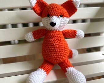 May include: A crocheted orange fox toy with white accents on the ears, paws, and nose. The toy is sitting on a white wooden bench.