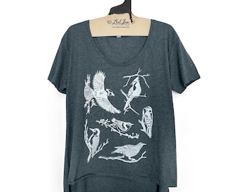 Könnte beinhalten: Ein dunkelgraues T-Shirt mit einem weißen Aufdruck von verschiedenen Vögeln auf Ästen. Das Shirt hat einen Rundhalsausschnitt und kurze Ärmel. Das Shirt ist locker geschnitten und hat eine längere Länge im Rücken.