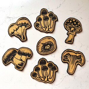 May include: A collection of seven mushroom-shaped cutouts. The cutouts feature a black outline and a tan interior, with varying mushroom cap designs. The mushrooms are arranged on a white, marbled surface.