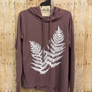 May include: A mauve long-sleeve hooded shirt featuring a white fern leaf graphic print on the front. The garment includes a hood with matching mauve drawstrings and a tag near the neckline that reads Mad Love and MadLoveShop.com.