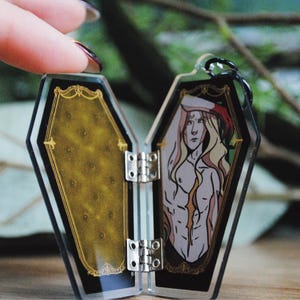 May include: A coffin-shaped pendant with a black and gold frame, hinged to open. Inside is an illustration of a shirtless figure with long blonde hair and a red hat. The other side has a gold patterned design. The pendant is held open.