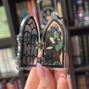 Könnte beinhalten: Ein durchsichtiger Acryl-Anhänger in Form eines gotischen Bogenfensters. Der Anhänger lässt sich aufklappen und zeigt eine Illustration von zwei sich umarmenden Fantasy-Figuren mit grüner und blauer Haut, umrahmt von schwarzen Fensterrahmen.