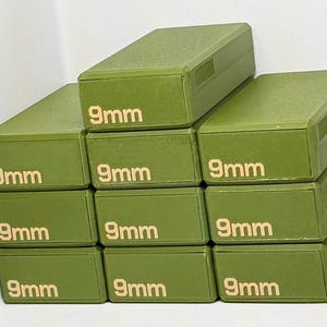 May include: A stack of olive green rectangular boxes. Each box has the text "9mm" printed in a gold color. The boxes are stacked in a staggered arrangement.