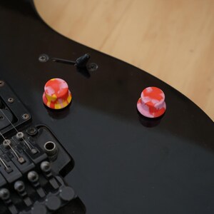 May include: Two colourful guitar knobs, one red and pink with white swirls, the other yellow, red, and white with a swirl pattern. The knobs are on a black electric guitar.