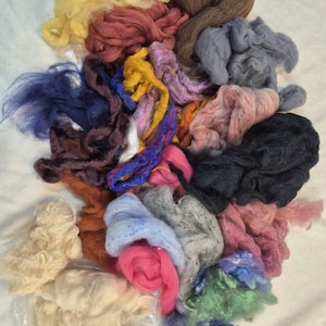 May include: A collection of raw wool roving in various colors including yellow, brown, blue, pink, black, and green. The fibers are arranged in loose, tangled clumps, showcasing different textures and shades of dyed and natural wool.