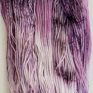 May include: A skein of hand-dyed yarn in shades of purple and white. The yarn is made of a soft, natural fiber and has a subtle ombre effect.