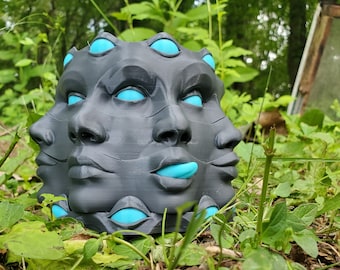 May include: A black and gray 3D printed planter shaped like a face with multiple eyes and mouths. The eyes and mouths are painted a bright turquoise color.