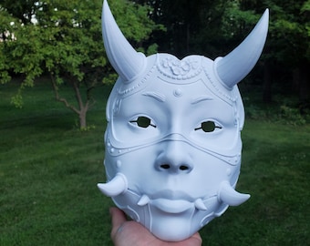 May include: White plastic mask with horns, intricate details, and a feminine face. The mask has a detailed design around the eyes and mouth.
