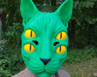 May include: A bright green cat mask featuring four distinct yellow eyes with vertical black pupils. The mask has pointed ears, a textured surface mimicking fur, and a small black nose. The design covers the entire face with symmetrical features.