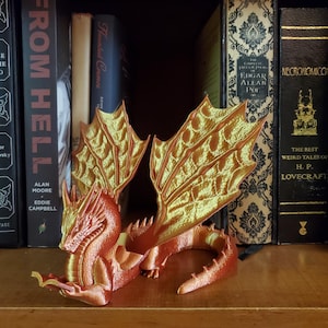 May include: A red and gold dragon figurine reading a book. The dragon has large wings and is posed in a sitting position. The dragon is on a shelf with books behind it.