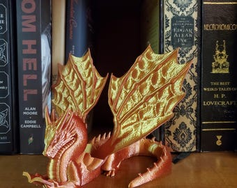 May include: A red and gold dragon figurine reading a book. The dragon has large wings and is posed in a sitting position. The dragon is on a shelf with books behind it.