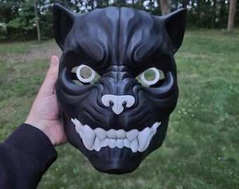 May include: A black, menacing mask with a snarling canine-like face. The mask features white teeth, a white nose, and circular eye openings. The mask is held up against a background of green grass and trees.